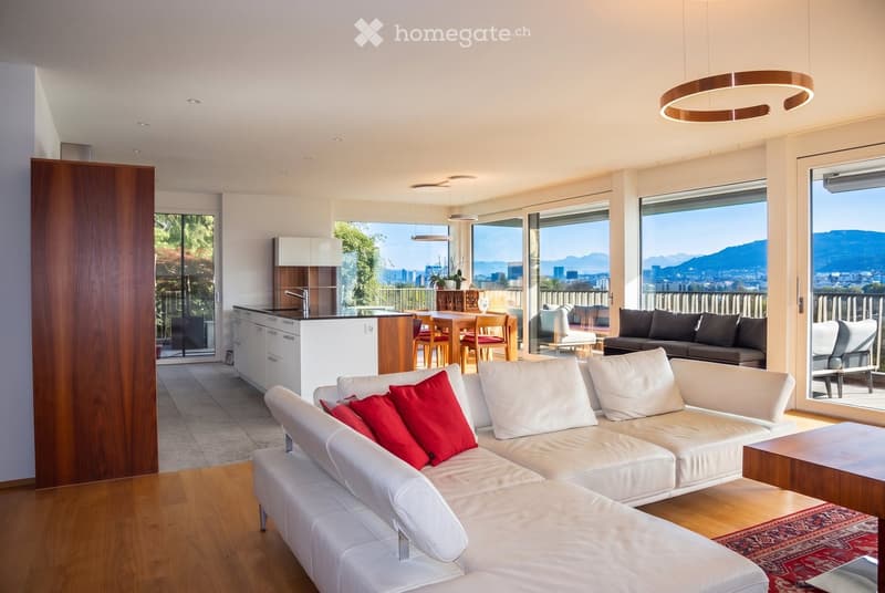 Modern living and dining area with white leather sofa, red throw pillows, coffee table, dining table with chairs, kitchen area, large windows offering a scenic view, and pendant lighting.