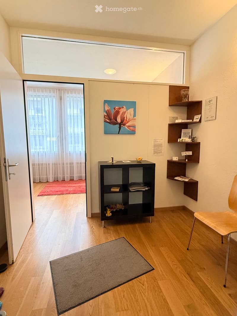 A small, well-lit room with wooden flooring, a shelf unit, a chair, a small table, a painting, and a curtained window leading to another space.