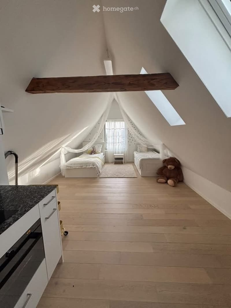 Loft-style bedroom with wooden floors, two single beds, nightstands, a small stool, and a kitchen area with an oven and faucet.