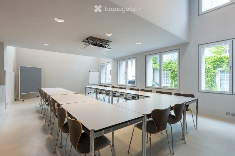 Bright, modern meeting room with white walls, large windows, and a long table surrounded by chairs. A projector screen and whiteboard are visible.