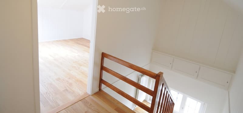 Empty room with light wooden flooring and white walls, featuring a wooden staircase railing and a visible doorway leading to another space.