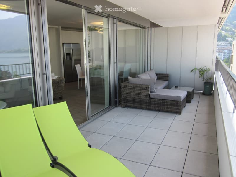 Balcony with tiled floor, modern furniture including a sofa, chairs, and a table, with a scenic view of mountains and water through large glass doors.