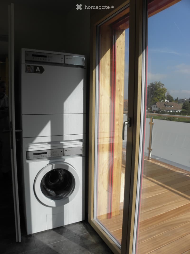 Modern bathroom with a washing machine and dishwasher stacked vertically, large window with view, wooden floor, and white tiled walls.