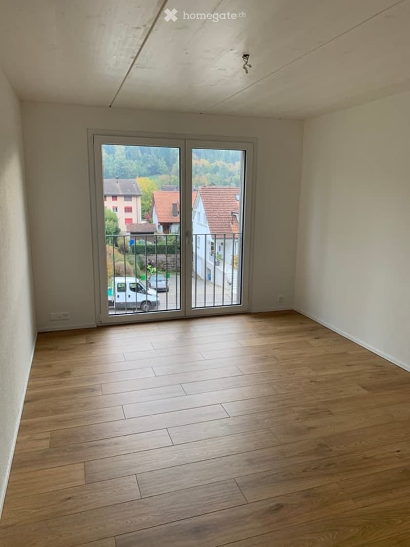 Empty room with wooden flooring and large sliding glass doors leading to a balcony or outdoor area. The walls are plain and there are no furnishings or decorations visible.