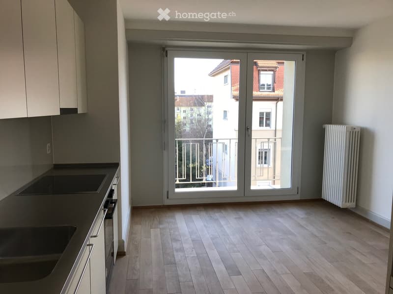 Empty kitchen with wooden flooring, white cabinets, a black countertop, and a large sliding glass door leading to a balcony with a view of neighboring buildings.