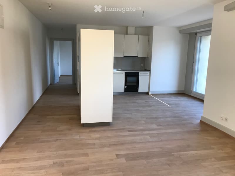 Empty room with wooden flooring, white walls, and a partially visible kitchen area with cabinets and appliances.