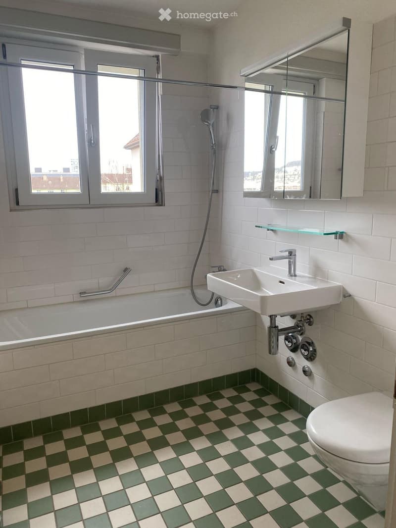 Modern bathroom with white tiles, green and beige checkered floor, bathtub, sink, mirror, and toilet.