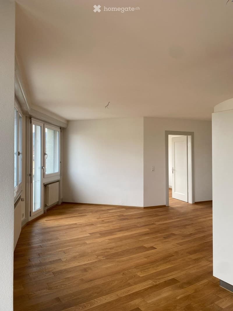 Empty room with wooden flooring, white walls, and large windows with sliding doors. A radiator is visible near the window, and an open doorway leads to another room.