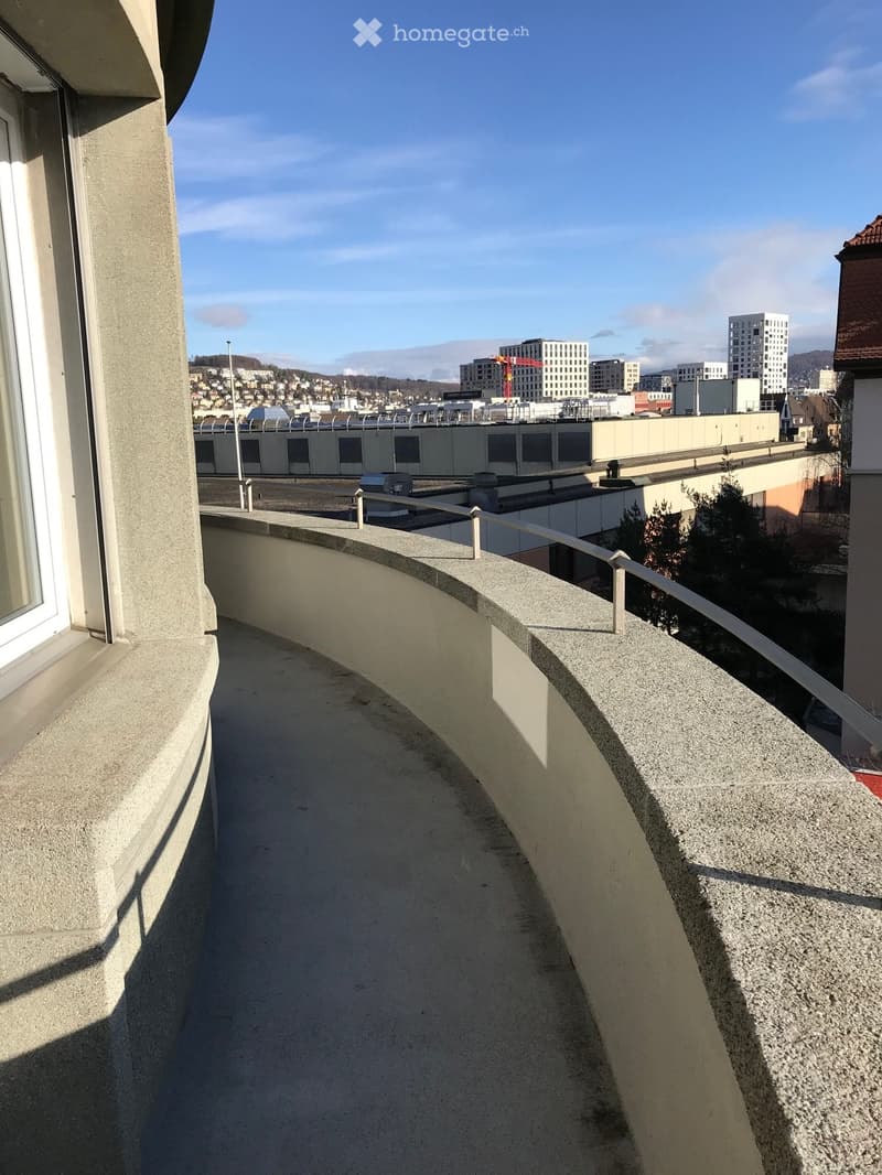 Balcony with concrete floor and railing, overlooking a cityscape with buildings and a clear sky.