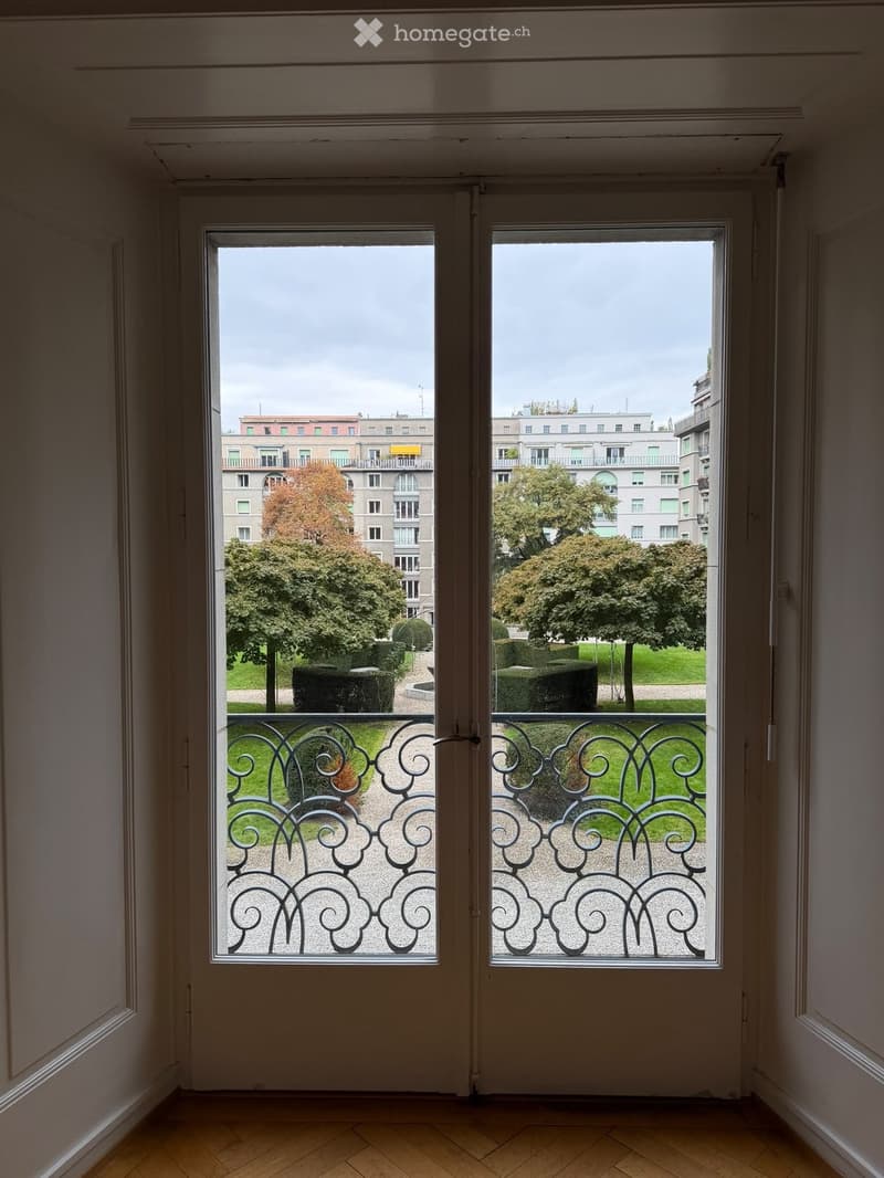 View from a room through a double door onto a balcony with ornate metal railings, overlooking a park with trees and buildings in the background.