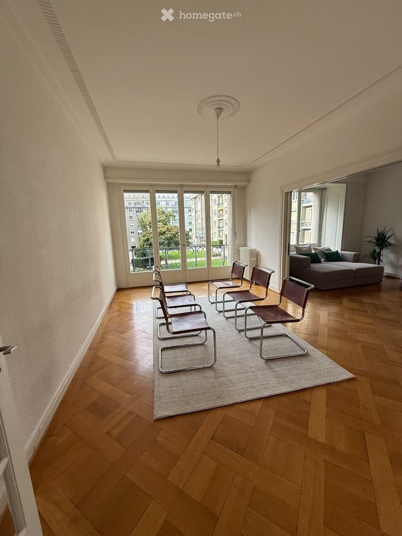 A modern living room with wooden flooring, a grey sofa, several chairs, and large windows offering a view of the city.