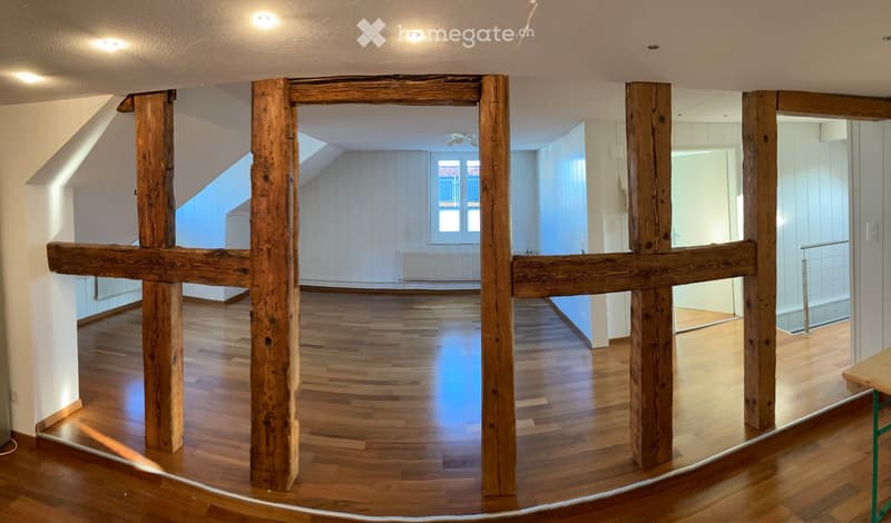 An empty, open-plan room with wooden beams and polished wooden flooring, featuring large windows and a modern design.