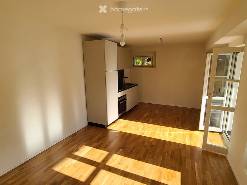Empty room with light wooden flooring, white walls, and a small kitchen area with cabinets and appliances visible in the background. Sunlight streams in from a window on the right side.