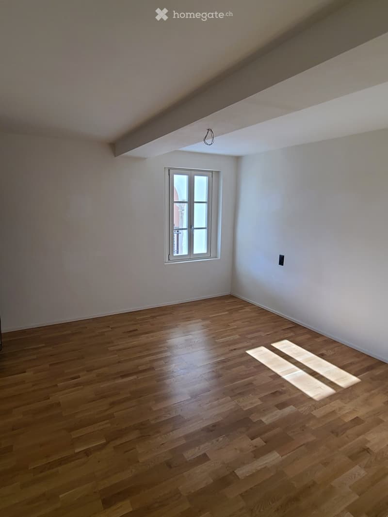 Empty room with wooden flooring, white walls, and a single window with white frame. The ceiling has a skylight and a hanging light fixture.
