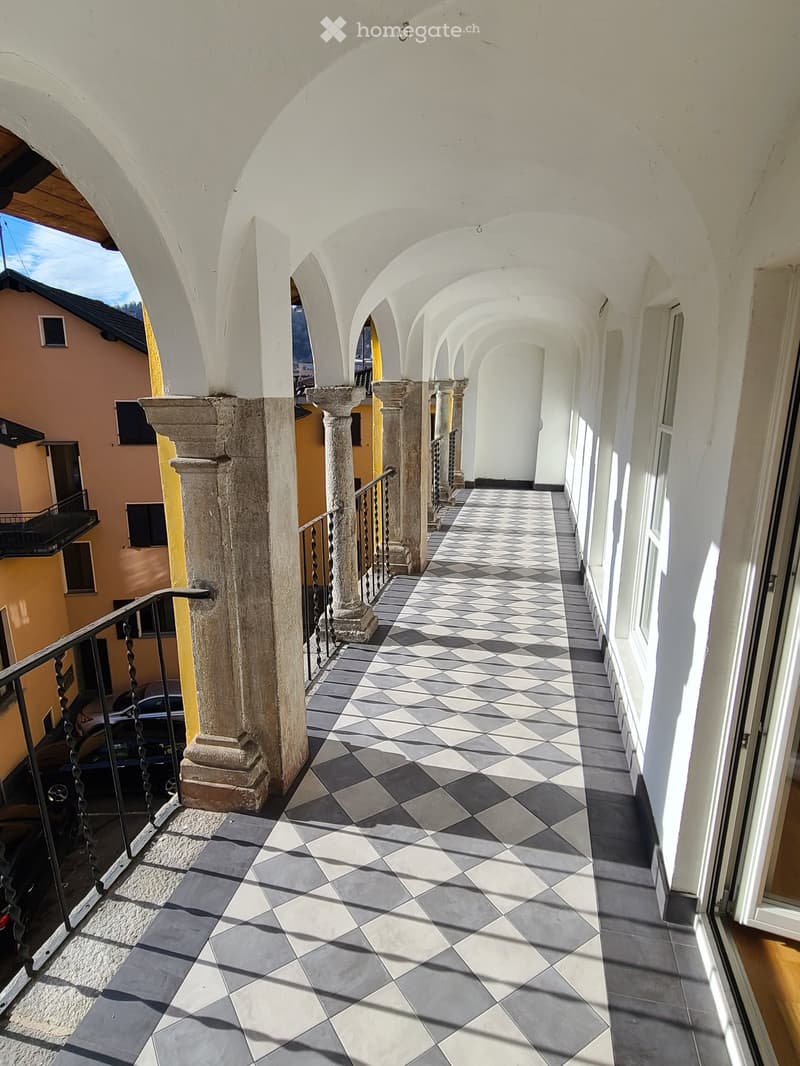 Arched corridor with checkered tile floor, stone columns, and metal railing leading to an open area.