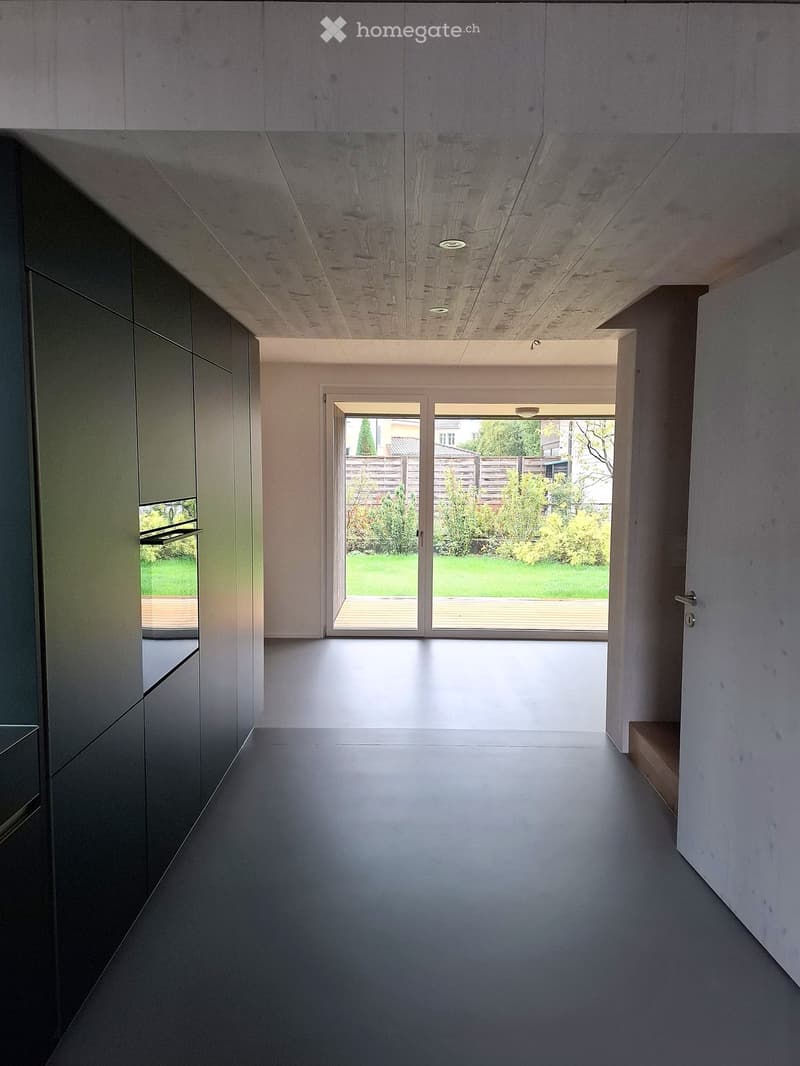 Modern, minimalist hallway with grey walls, dark grey floor, and a large glass door leading to a garden area.