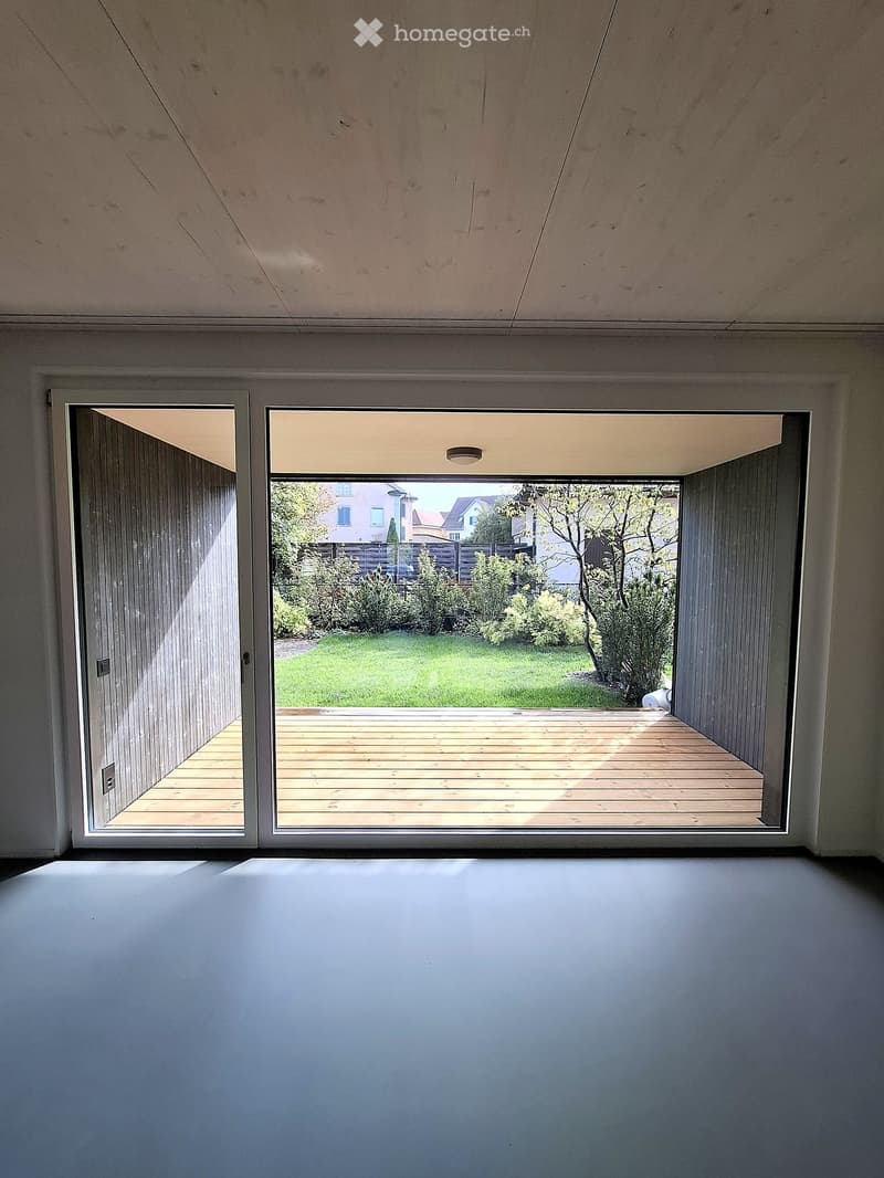Empty room with large sliding glass doors leading to a wooden deck and a green garden area.