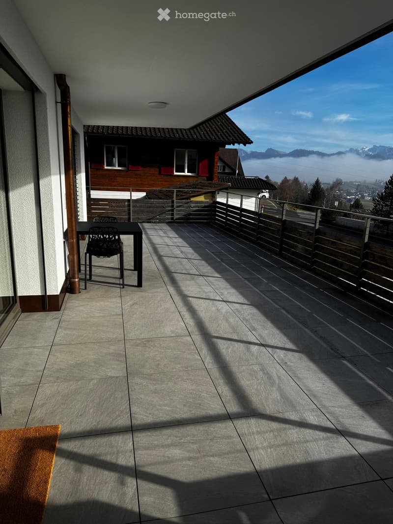 A tiled terrace with a black table and chairs, surrounded by a wooden fence, offering a scenic view of mountains and a town.