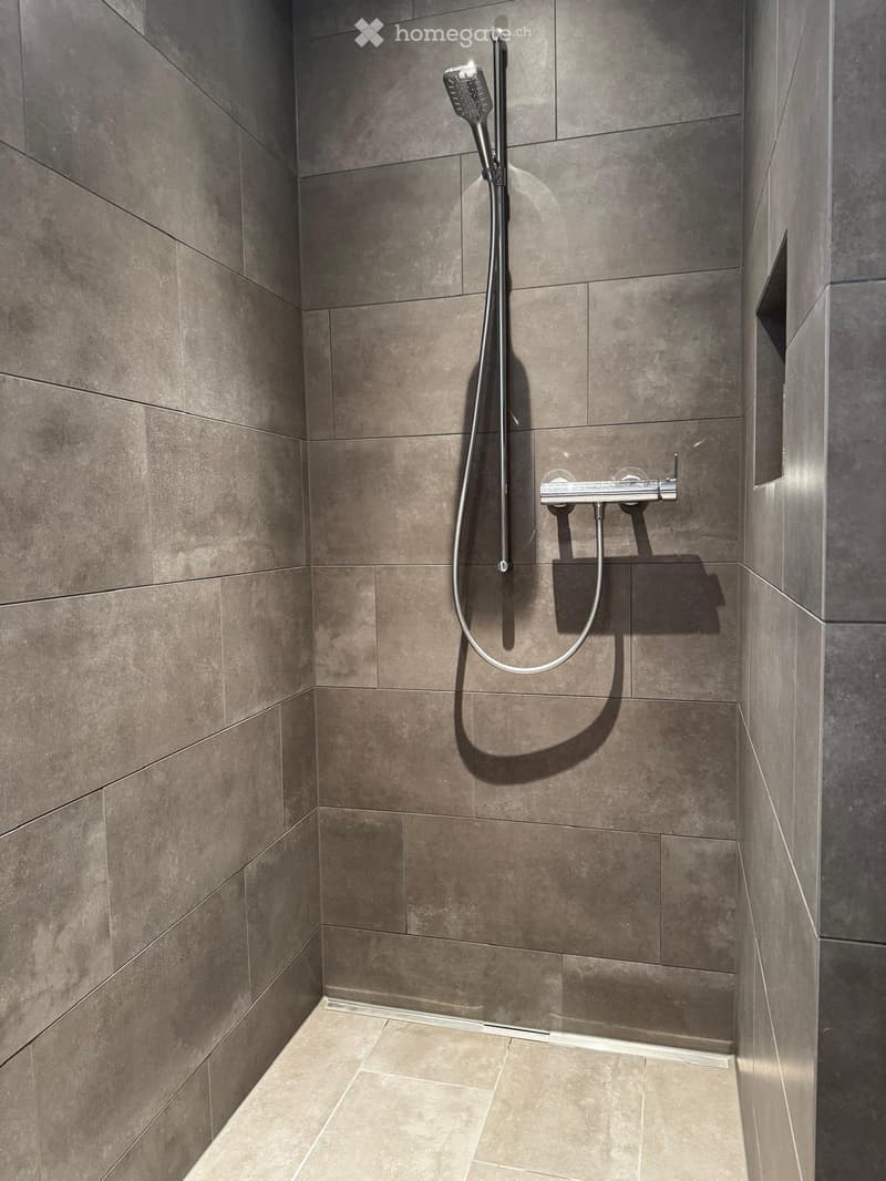The shower area is fully tiled with light grey tiles, drain channel installed in the floor, a handheld shower head attached to a shower arm.