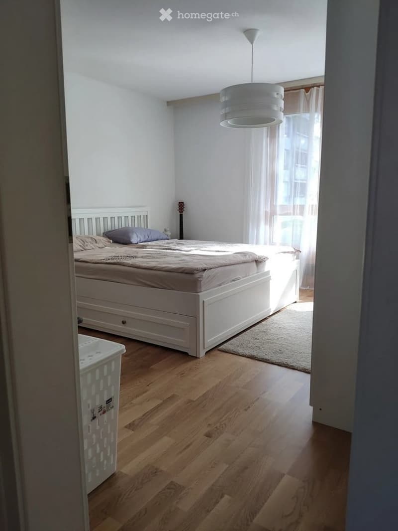 Bright bedroom with wooden flooring, white bed with beige bedding, a guitar leaning against the wall, a small rug, and a laundry basket near the door.
