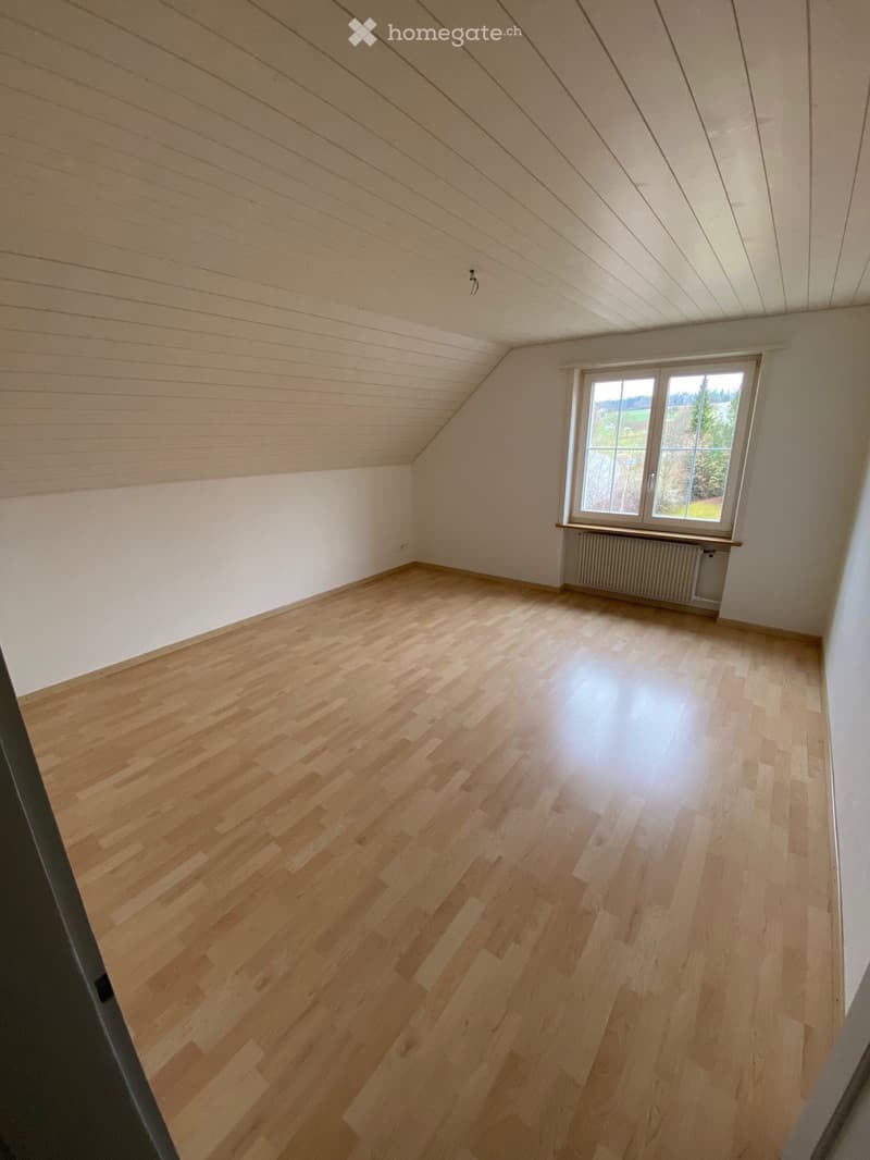 Empty room with wooden flooring, white walls, and a large window with a view of greenery. The ceiling has a sloped design with visible beams.