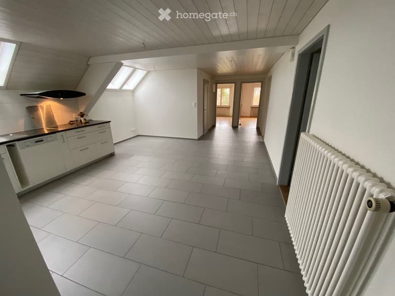 Empty hallway with tiled floor, white walls, and a radiator on the right side. The hallway leads to a kitchen area with white cabinets and a countertop on the left side.