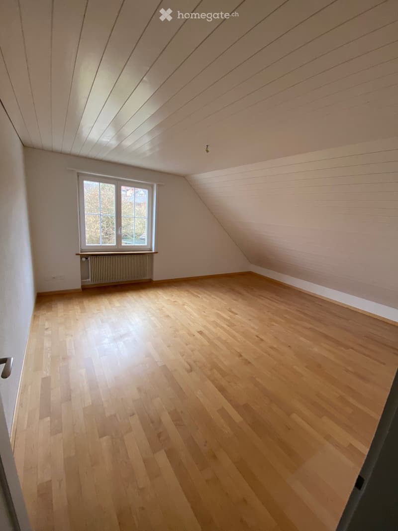Empty attic room with wooden flooring, slanted ceiling, and a single window with a radiator below it.