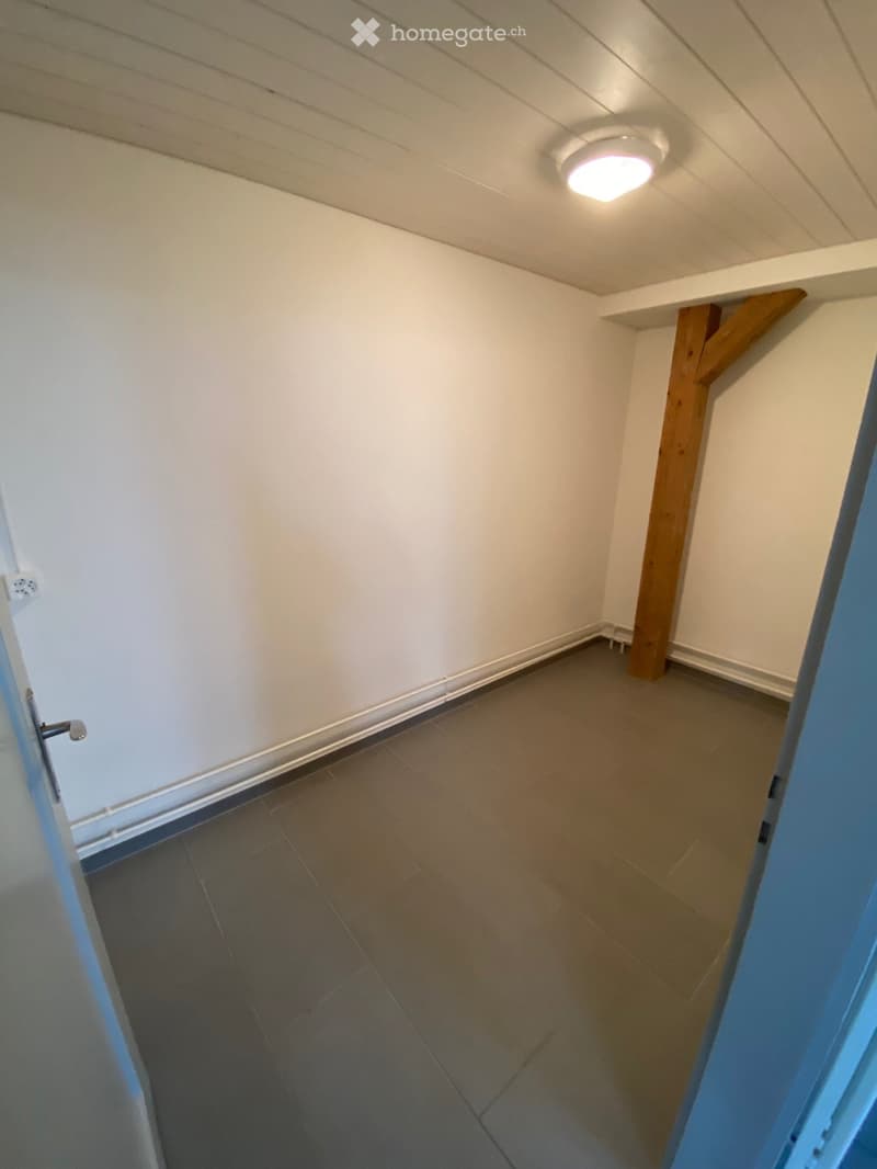 Empty room with tiled floor, white walls, and a wooden support beam. A ceiling light fixture is visible.