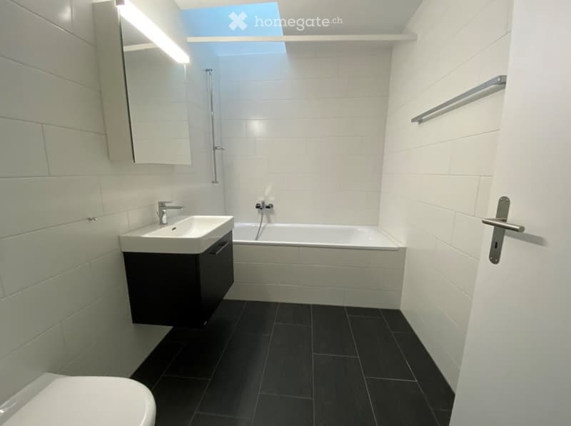 Modern bathroom with white tiles, black floor, sink, bathtub, toilet, and mirror.