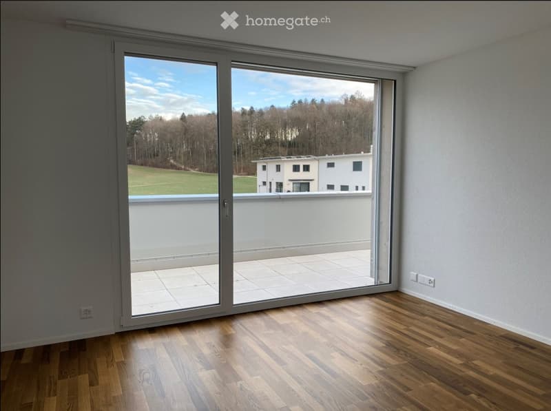 Empty room with wooden flooring, large sliding glass doors leading to a balcony, and a scenic view of a green field and trees.