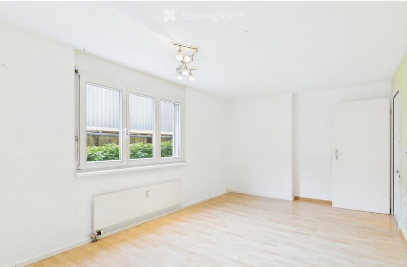 Empty room with white walls, wooden flooring, large windows, radiator, and a ceiling light fixture.
