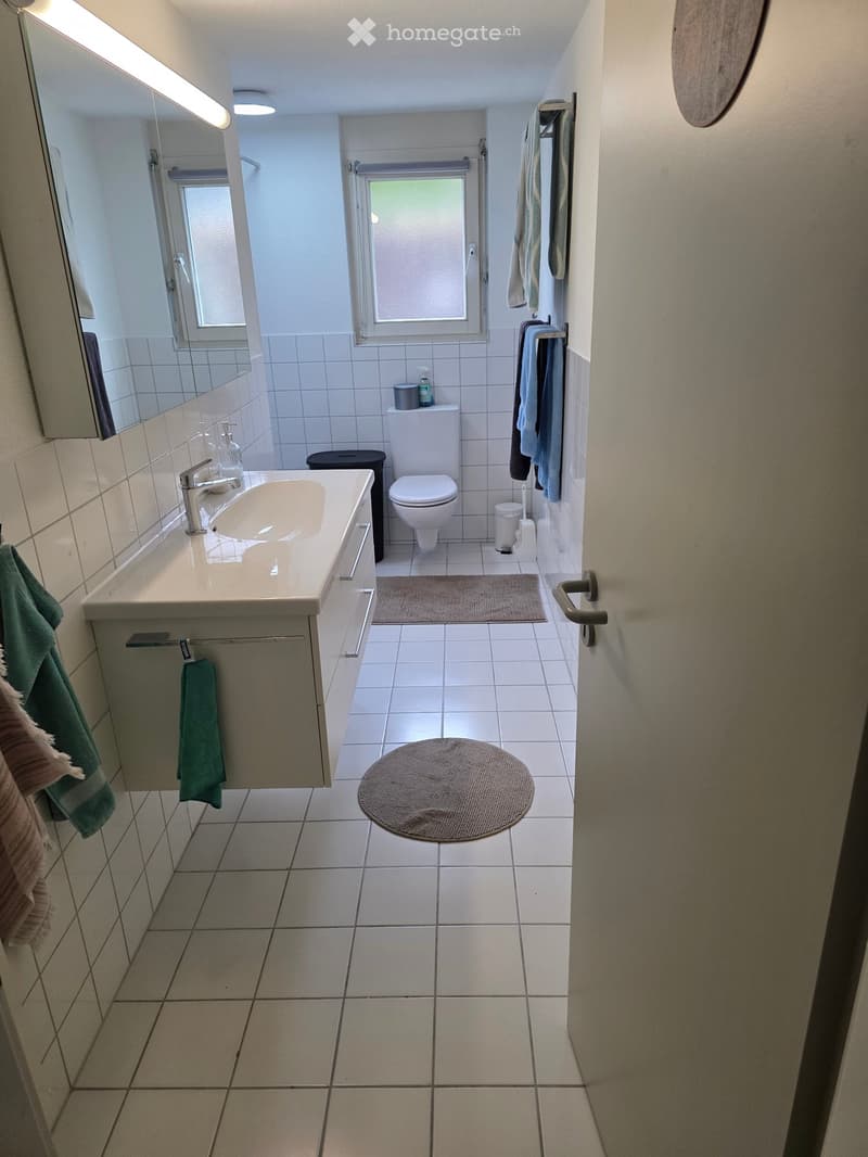 Modern bathroom with white tiles, a sink, toilet, mirror, and towels hanging on the wall.