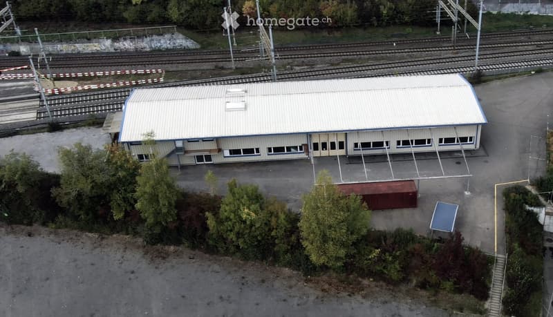 A single-story industrial building with a metal roof and multiple windows, situated near railway tracks. The surrounding area includes trees, a small container, and a blue structure.