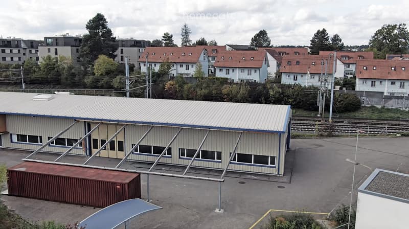 A modern industrial-style building with a metal roof and large windows, situated in an urban area with surrounding residential houses and trees.