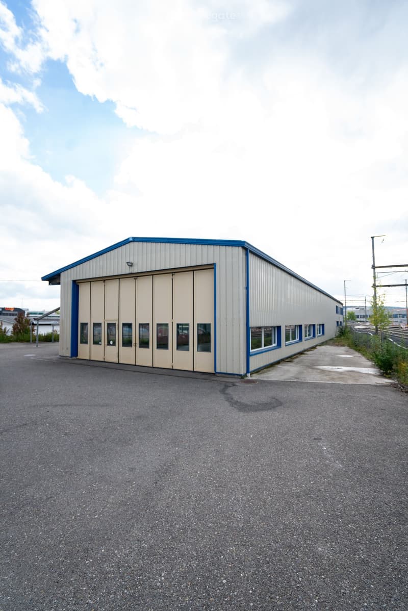 A single-story industrial building with a large garage door and multiple windows, surrounded by a paved area.