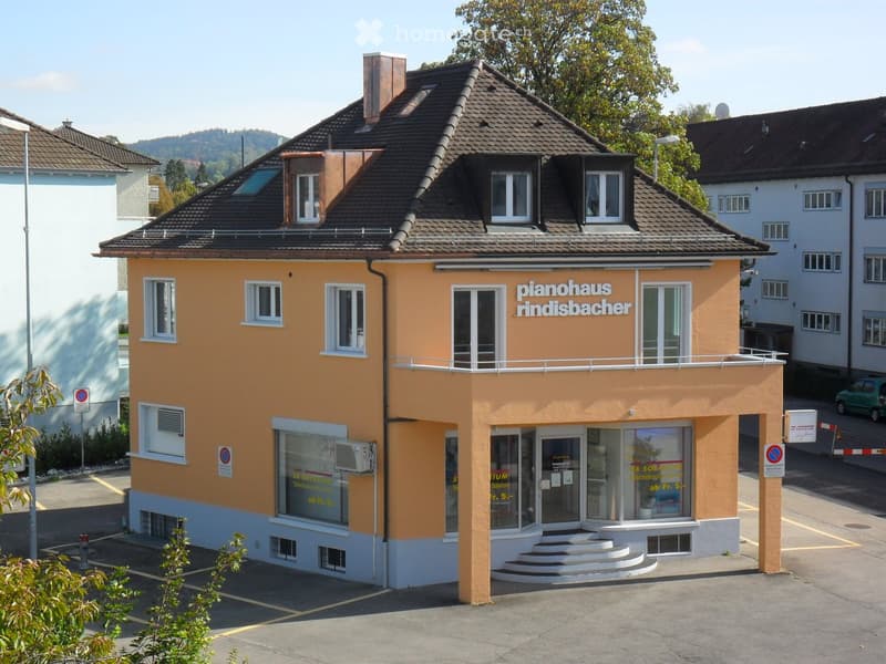 Two-story building with a brown roof, yellow walls, white windows, and a balcony on the second floor. The building has a sign that reads 'pianohaus rindisbacher'. There is a parking lot in front of the building, and a green car is parked on the side of th
