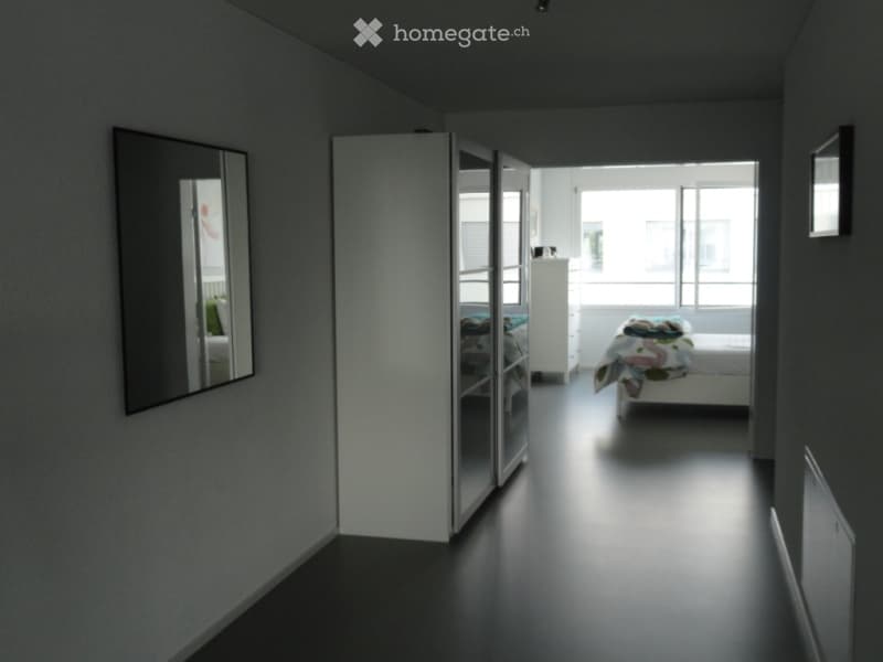 Modern, minimalist bedroom with dark flooring, large mirror, sliding wardrobe, and neatly made bed.
