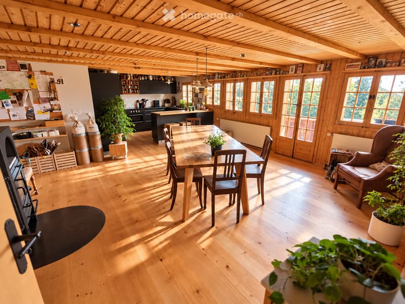 Bright, open-plan living and dining area with wooden floors, exposed wooden beams, large windows, a dining table with chairs, a sofa, and a kitchen area with cabinets and appliances.