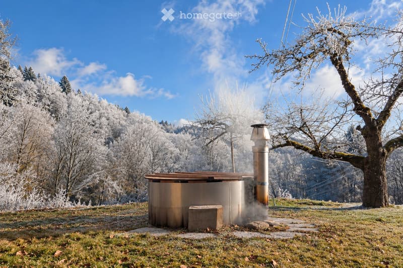 A wooden hot tub with a metal chimney is situated in a grassy area surrounded by frost-covered trees and a clear blue sky.