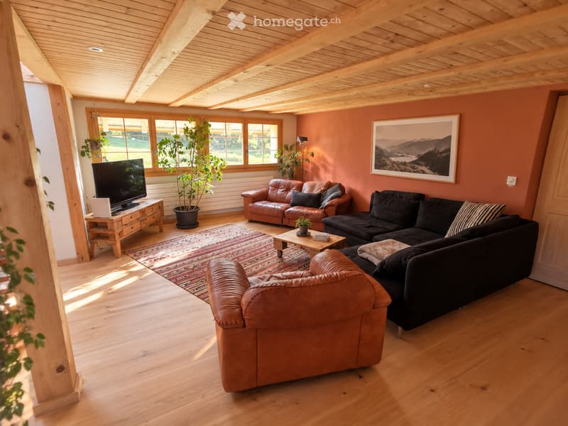 Bright living room with wooden beams, hardwood floors, and large windows. Features a black sectional sofa, brown leather armchair, coffee table, TV on a wooden stand, and a potted plant.