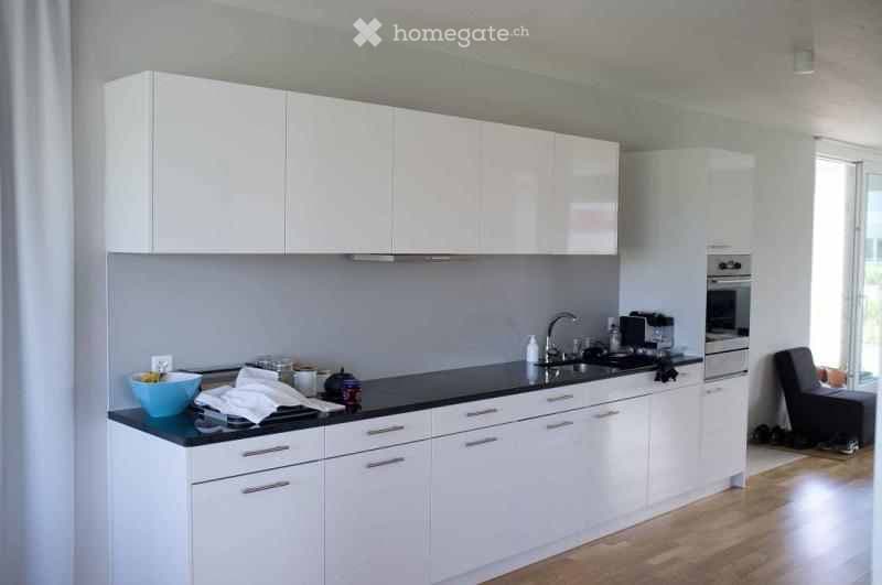 Modern kitchen with white cabinets, black countertop, and stainless steel appliances. Features a sink, oven, and a small seating area with a stool.