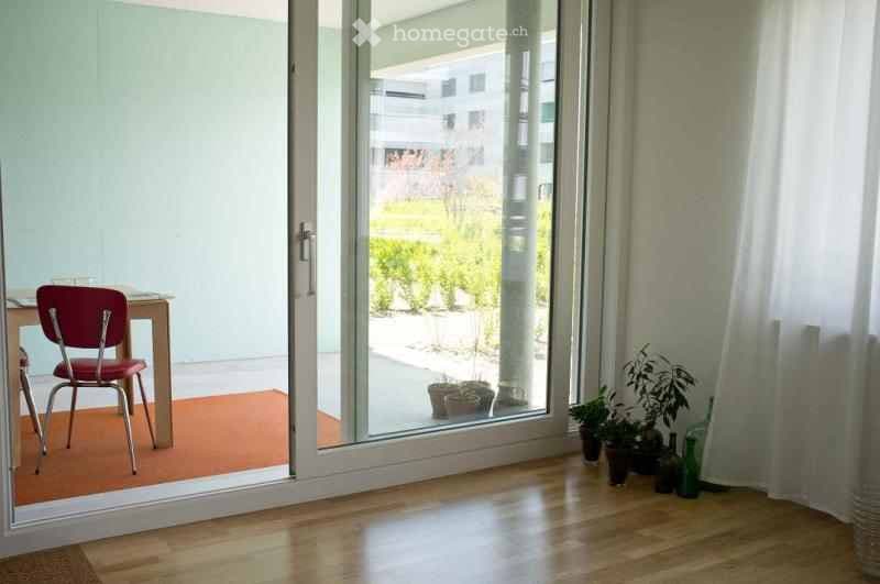A small, well-lit room with wooden flooring, a red chair, a small table, and several potted plants near the sliding glass door that opens to a garden area.