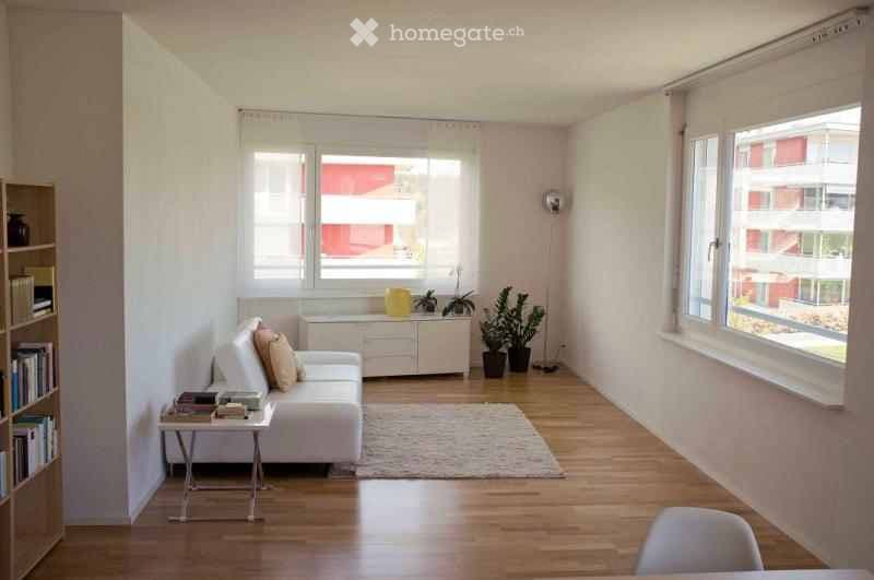 Bright, modern living room with white walls, wooden flooring, large windows, a white sofa with cushions, a coffee table, a bookshelf, and potted plants.