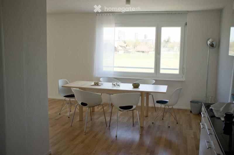 Bright dining area with a wooden table and white chairs, large windows with sheer curtains, and a modern floor lamp near the wall.