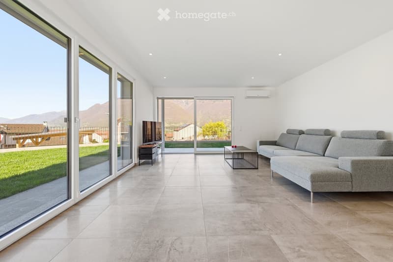 Modern living room with large windows offering a scenic mountain view, featuring a grey sectional sofa, a coffee table, a television, and a small side table.
