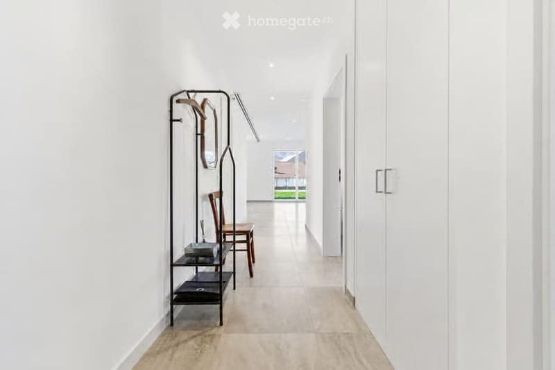 Modern, minimalist hallway with white walls, light wooden flooring, and built-in storage units. A coat rack and a small chair are visible on the left side.