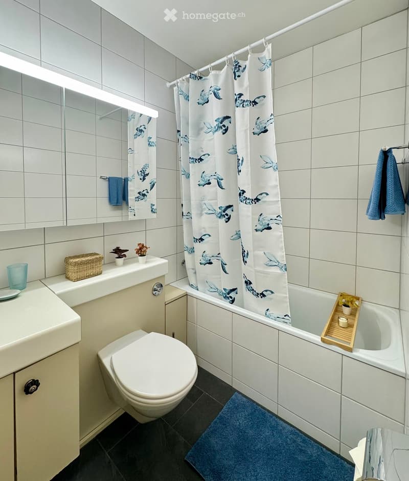 Modern bathroom with white tiles, a bathtub with a decorative shower curtain, a toilet, a sink with a mirror, and small decorative items on the counter.