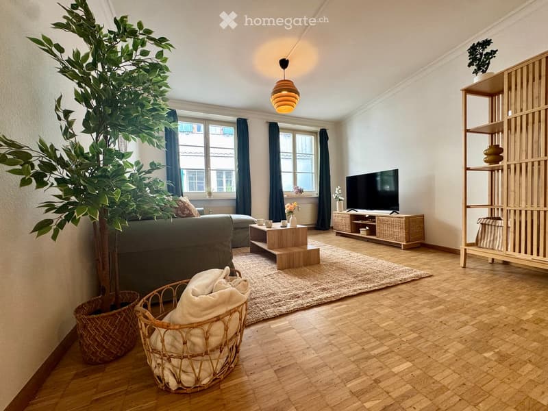 Modern living room with wooden flooring, large windows, a grey sofa, coffee table, TV, floor lamp, and decorative shelves.
