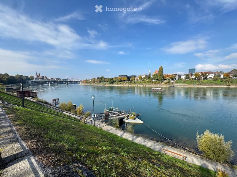 Riverfront view with a bridge, boats, and cityscape in the background. Pathway with benches and greenery along the riverbank.