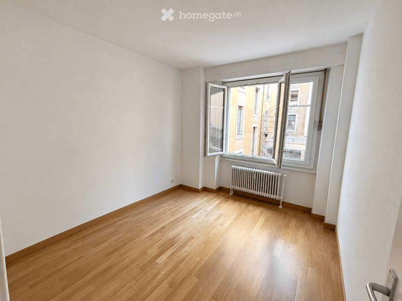 Empty room with wooden flooring, white walls, and a radiator under a large window.
